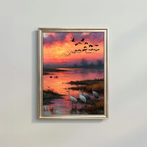 May include: A framed landscape painting depicting a vibrant sunset over a marshland with a flock of birds flying in the sky and several white cranes standing in the shallow water, rendered in shades of orange, pink, purple, and deep blue.