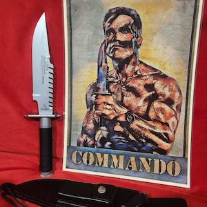 May include: A large survival knife with a serrated blade and black handle, next to a framed print of a man holding a knife, with the word "COMMANDO" at the bottom. A black leather sheath is also present.