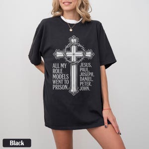 May include: Black t-shirt featuring a large, detailed cross design with a crown of thorns and the text "ALL MY ROLE MODELS WENT TO PRISON." The names "JESUS, PAUL, JOSEPH, DANIEL, PETER, JOHN" are also listed.