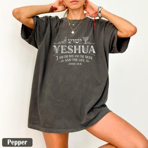 May include: A dark grey t-shirt featuring the word "YESHUA" in white, with the text "I AM THE WAY AND THE TRUTH AND THE LIFE - JOHN 14:6". The shirt also includes Hebrew text and a decorative design. The shirt is labelled "Pepper" at the bottom.