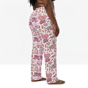 May include: White pajama pants with a repeating pattern of shopping bags, dollar signs, and the text "Spend Dat Shit" in pink and purple graffiti style. The pants have a relaxed fit.