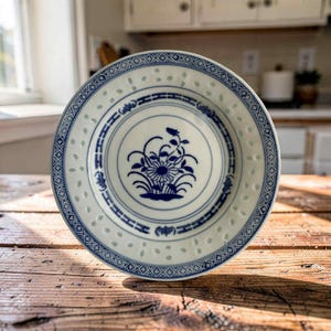 May include: A round ceramic bowl featuring a white base with blue floral patterns in the centre. The rim displays a geometric border and a series of small, translucent cut-out holes. The bowl has a smooth, glazed finish and a circular shape.
