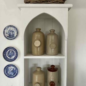 May include: A white wooden shelving unit with an arched top, holding four cylindrical stoneware bottles with side spouts. A woven wicker basket sits on top. Three blue and white decorative plates are mounted on the wall to the left of the unit.