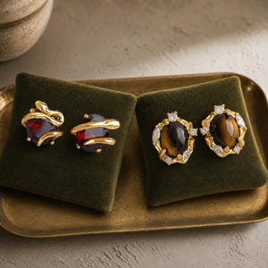May include: Two pairs of gold-tone earrings. The first pair features deep red oval stones wrapped in gold snake motifs. The second pair showcases oval tiger's eye stones surrounded by gold settings accented with small, clear, sparkling stones.