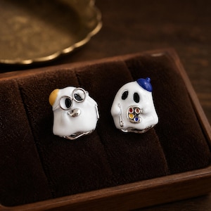 Può includere: Due spille in smalto a forma di fantasma bianco. Il fantasma a sinistra indossa occhiali tondi con montatura argentata e ha una macchia gialla. Il fantasma a destra indossa un basco blu e tiene in mano un pennello e una tavolozza.