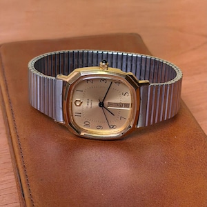 May include: A gold-toned Timex Quartz watch with a rectangular face and a silver expansion band. The watch face displays the time with black hands and numerals. The watch is resting on a brown leather surface.
