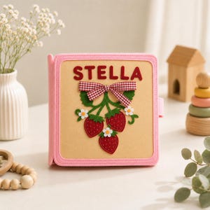 May include: A square felt book with a light pink border and a tan cover. The cover features the name STELLA in red letters, a red and white gingham ribbon bow, and three red strawberries with green leaves and small white flowers with yellow centers.