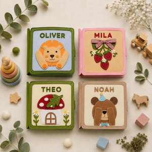May include: Four square felt books with embroidered names. Top left: Oliver with a lion. Top right: Mila with strawberries and a red gingham bow. Bottom left: Theo with a mushroom house. Bottom right: Noah with a bear wearing a blue party hat.