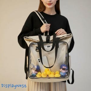 Könnte beinhalten: Ein beigefarbener Rucksack aus Segeltuch mit einer großen, durchsichtigen Kunststofftasche auf der Vorderseite. Der Rucksack hat schwarze Stoffriemen und einen Schnallenverschluss. Im Inneren befinden sich ein weißes Smartphone und gelbe Enten.