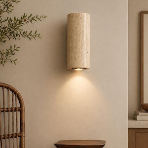 May include: A cylindrical wall sconce made of beige stone is mounted on a light-colored wall. The sconce emits a warm light downwards. The image also shows a wooden side table, a wicker chair, and a framed artwork.