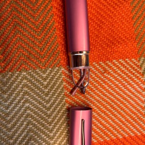 May include: A pink, cylindrical glasses case with a silver accent and a matching pair of reading glasses. The case is open, revealing the glasses. The background is a textured orange and brown fabric.