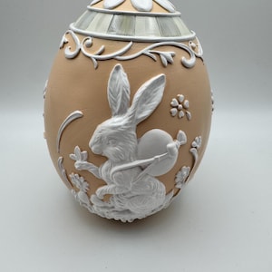 May include: A decorative egg-shaped container with a tan base and white floral and bunny relief details. The lid is white with a floral design. The bunny is holding a round object. The egg is a decorative item.