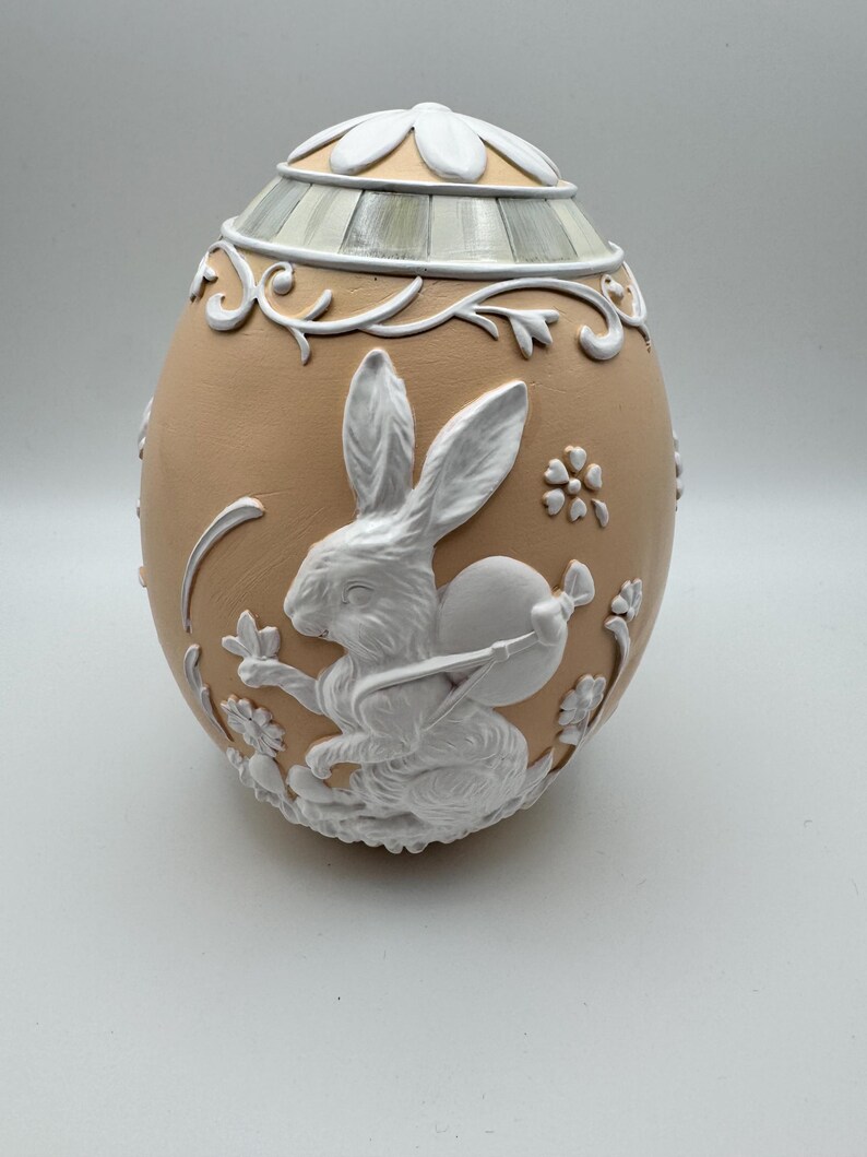 May include: A decorative egg-shaped container with a tan base and white floral and bunny relief details. The lid is white with a floral design. The bunny is holding a round object. The egg is a decorative item.