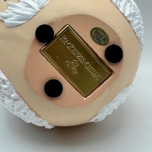 May include: Close-up of the bottom of a decorative figurine. The base is a light peach color with three black circular feet. A gold rectangular plaque reads "MacKenzie-Childs" and "Made in China" is printed on a circular sticker.