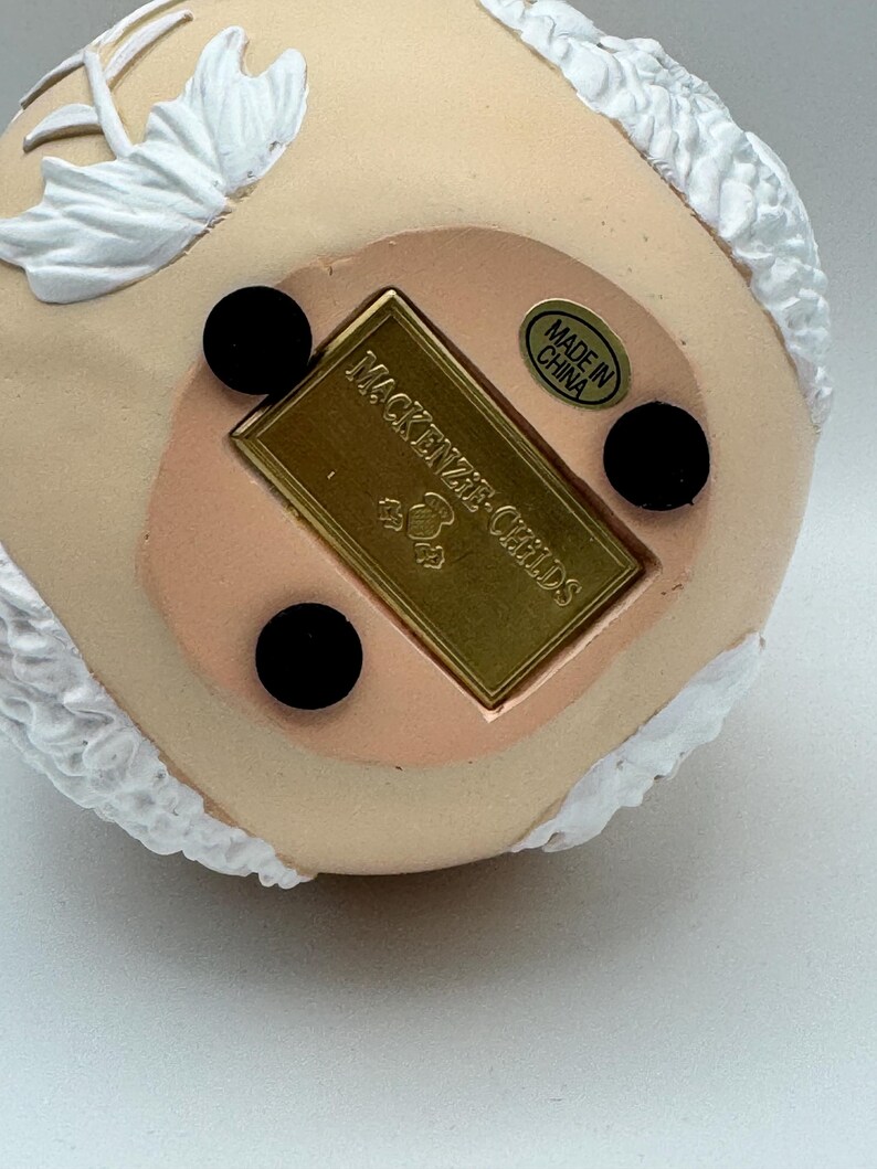 May include: Close-up of the bottom of a decorative figurine. The base is a light peach color with three black circular feet. A gold rectangular plaque reads "MacKenzie-Childs" and "Made in China" is printed on a circular sticker.