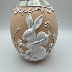May include: A decorative egg-shaped ornament in a light peach color, featuring a white relief design of a rabbit carrying a satchel. The egg has a floral and vine pattern, with a detailed lid. A charming Easter-themed decorative item.