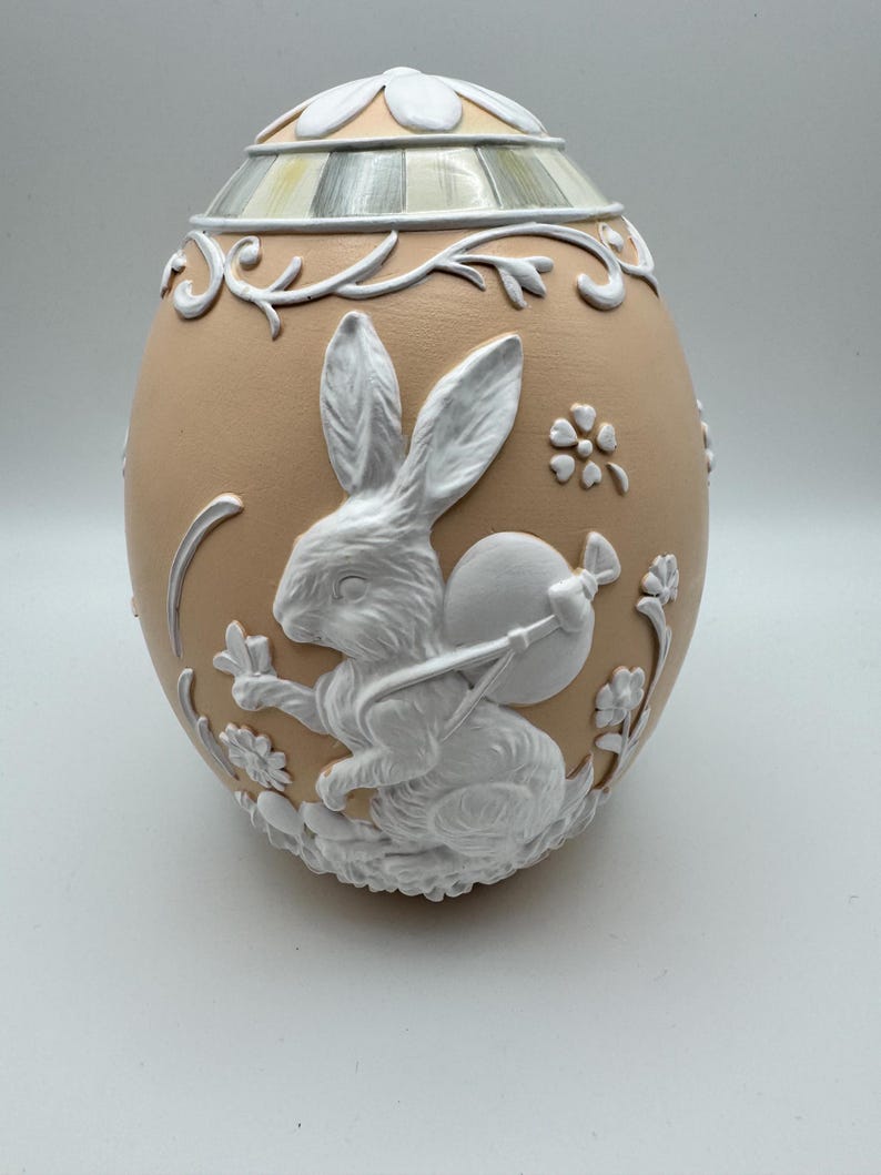 May include: A decorative egg-shaped ornament in a light peach color, featuring a white relief design of a rabbit carrying a satchel. The egg has a floral and vine pattern, with a detailed lid. A charming Easter-themed decorative item.