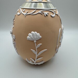 May include: A decorative egg-shaped container with a tan base and white floral and scrollwork accents. The top features a striped design and a white floral pattern. The egg is a decorative item.