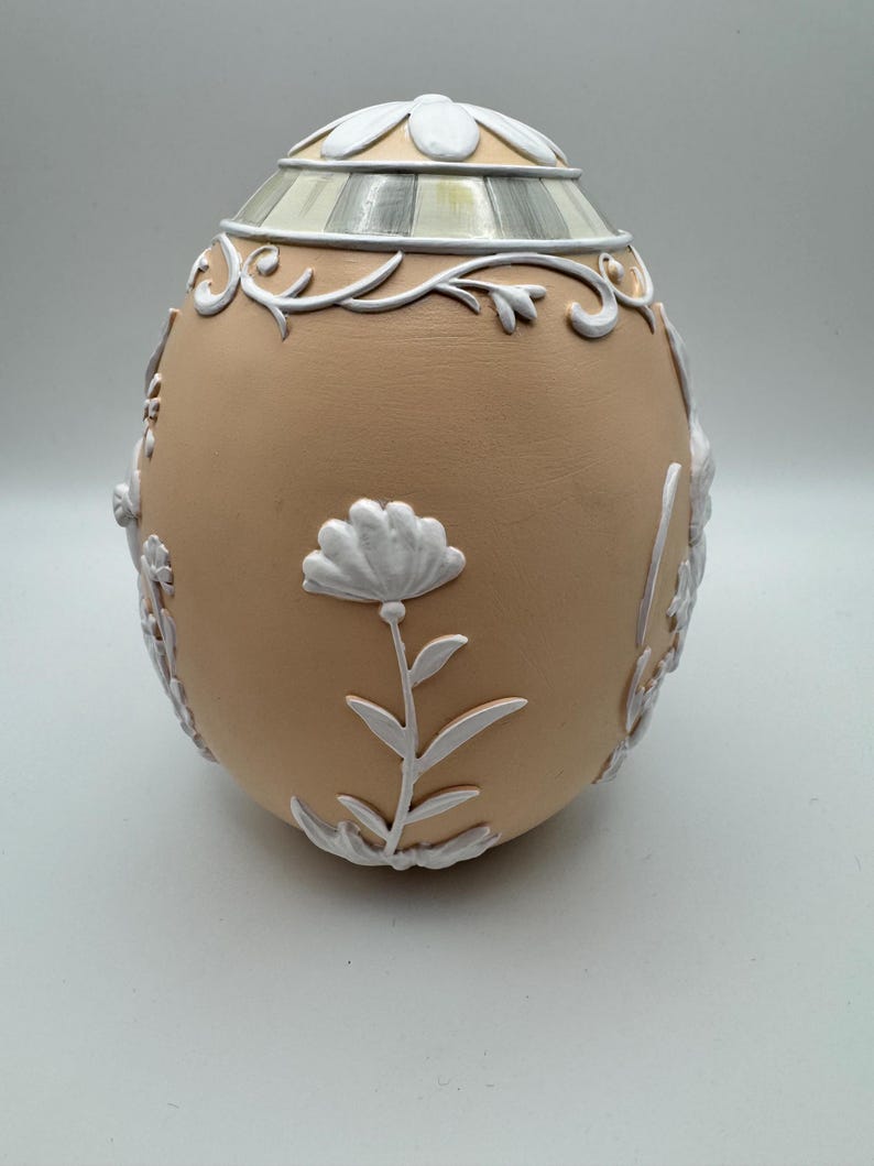 May include: A decorative egg-shaped container with a tan base and white floral and scrollwork accents. The top features a striped design and a white floral pattern. The egg is a decorative item.