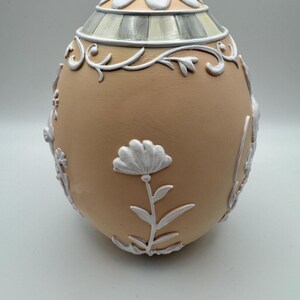 May include: A decorative egg-shaped container with a tan base and white floral and scrollwork accents. The top features a flower-shaped design and striped detailing. The egg is a unique decorative item.