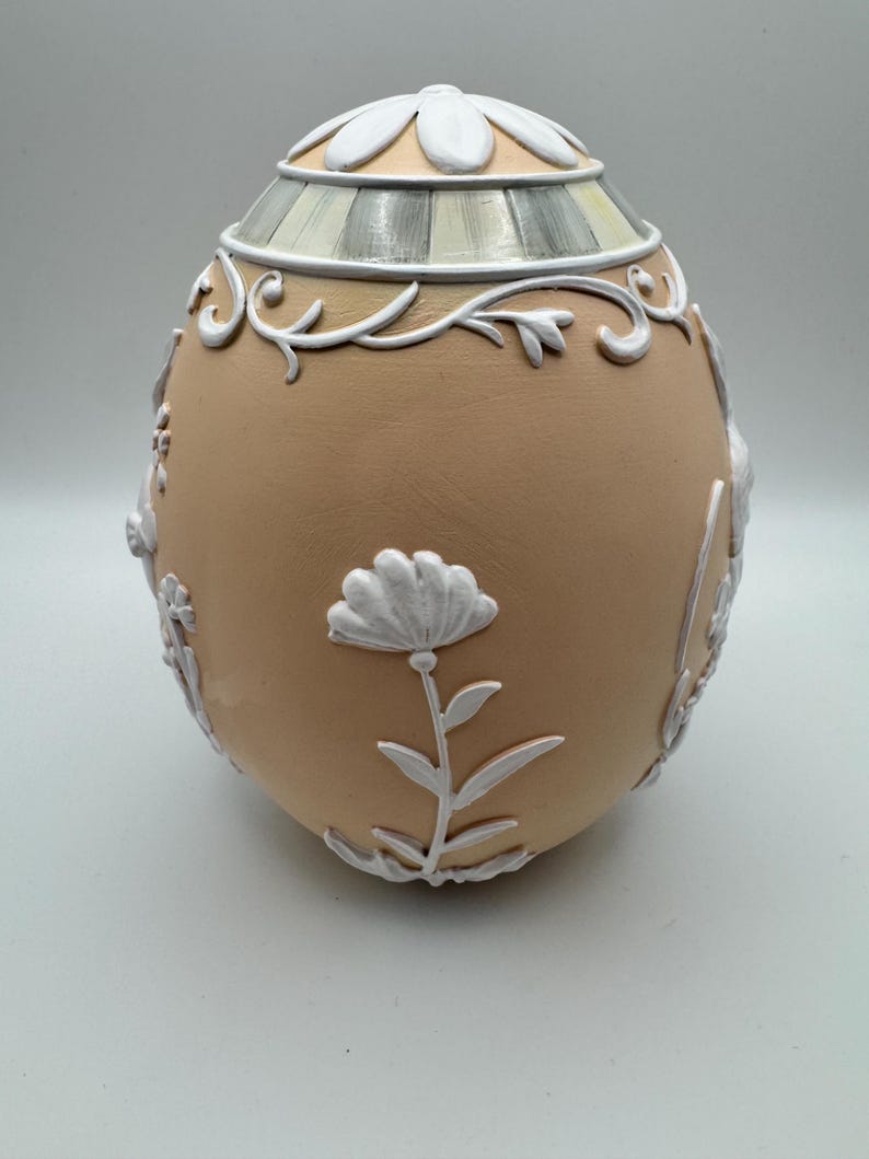May include: A decorative egg-shaped container with a tan base and white floral and scrollwork accents. The top features a flower-shaped design and striped detailing. The egg is a unique decorative item.
