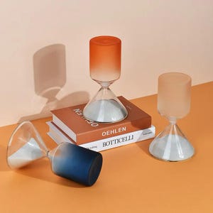 Peut inclure: Trois sabliers avec des parties supérieures en verre dépoli et des bases en verre transparent contenant du sable blanc. Un sablier est orange, un est pêche clair et un est bleu foncé. Deux sabliers sont debout, le bleu est couché.