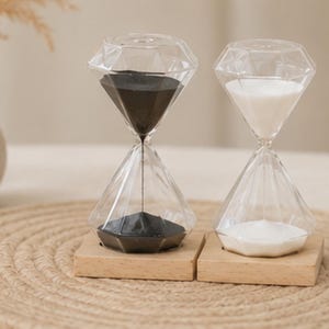 Peut inclure: Deux sabliers en verre en forme de diamant avec des bords facettés, chacun monté sur une base carrée en bois clair. Un sablier contient du sable noir fin, tandis que l'autre contient du sable blanc fin, s'écoulant par un centre étroit.