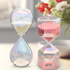 May include: Two hourglasses with iridescent glass frames. The left hourglass has a rounded shape and contains white sand. The right hourglass features a faceted hexagonal design and contains pink sand. Both items have a reflective, rainbow-like finish.
