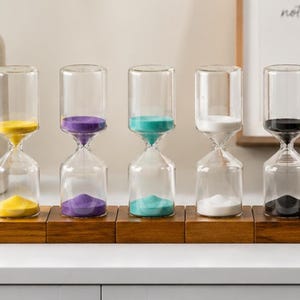 May include: Five glass hourglasses with colored sand (yellow, purple, teal, white, and black) sit on a wooden base. The hourglasses are clear, allowing the sand to be seen as it flows from top to bottom.