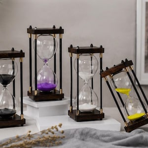 May include: Four hourglasses with dark wooden frames and clear glass bulbs. Each hourglass contains a different colour of sand: black, purple, white, and yellow. The hourglasses are arranged on a white surface with a grey cloth.