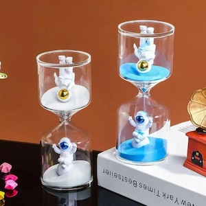 Puede incluir: Dos relojes de arena de cristal transparente con figuras de astronautas blancos en su interior. Uno contiene arena blanca y el otro arena azul. Cada astronauta tiene una visera dorada o azul. Los objetos están sobre un libro blanco.