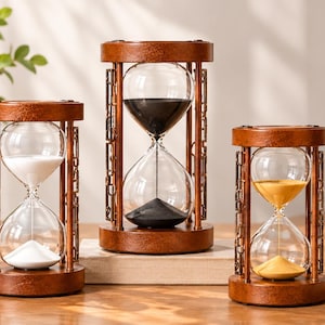 May include: Three hourglasses with brown wooden frames and intricate side supports. The left hourglass contains white sand, the centre one holds black sand, and the right one features yellow sand. Each glass bulb is clear and set within a circular base.