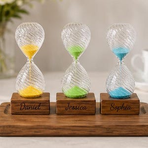 Op de afbeelding: Drie glazen zandlopers met gedraaide ribbelpatronen, elk gevuld met geel, groen of blauw zand. De zandlopers staan op individuele rechthoekige houten voetstukken die zijn gegraveerd met de namen Daniel, Jessica en Sophia.