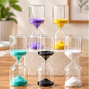 May include: A set of five clear glass hourglasses arranged in two rows. The hourglasses contain fine sand in distinct colors: purple, yellow, turquoise, black, and white. Each hourglass features a cylindrical glass frame with a narrow center.