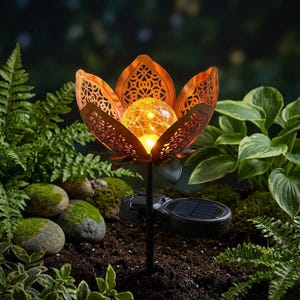 Op de afbeelding: Een op zonne-energie werkende tuinpaallamp in de vorm van een bloem. De koperkleurige bloemblaadjes hebben een decoratief uitgesneden ontwerp, dat een gloeiende glazen bol omringt. De paal is zwart en het zonnepaneel is aan de basis zichtbaar. De achtergrond bestaat uit groen gebladerte.