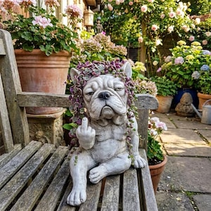 Puede incluir: Estatua de bulldog de hormigón con un gesto de dedo medio, adornada con una corona de plantas moradas. La estatua está sentada en un banco de madera en un jardín, rodeada de plantas y flores en macetas. El bulldog tiene una expresión gruñona.