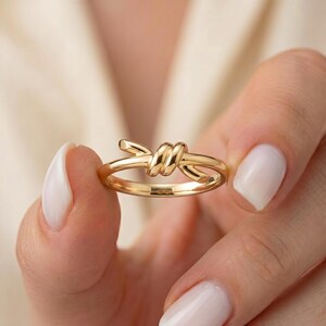 May include: A polished gold ring featuring a central knot design. The band is smooth and circular, while the focal point consists of two overlapping loops of metal that form a stylized knot, creating a continuous and elegant metallic silhouette.