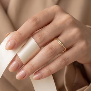 Puede incluir: Un anillo de oro pulido con un contorno suave y ondulado. La banda presenta un perfil redondeado y un acabado metálico de alto brillo, rodeando el dedo con una curva suave y sinuosa que crea una estética fluida y sencilla.