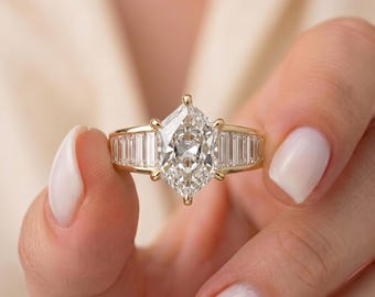 May include: A gold ring featuring a large, elongated hexagonal-cut center diamond held by six prongs. The band is accented with a row of channel-set baguette-cut diamonds on each side, creating a symmetrical and polished appearance in yellow gold.