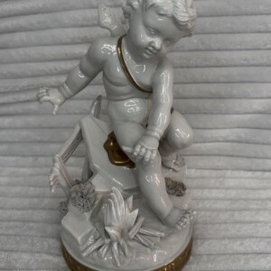 May include: A white porcelain cherub figurine, featuring a bow and arrow, seated on a decorative base. The cherub has a gold strap across its chest and is surrounded by floral and architectural elements. The base has a gold trim.