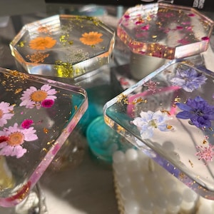 May include: Four clear resin coasters with dried flowers and gold flakes. The coasters are square and hexagonal, with pink, orange, and purple flowers embedded in the resin. A decorative item.