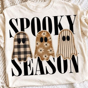 May include: A cream-colored t-shirt featuring the black text SPOOKY SEASON and three ghost illustrations. The first ghost has a black and cream buffalo check pattern, the second has a brown floral print, and the third has vertical stripes.