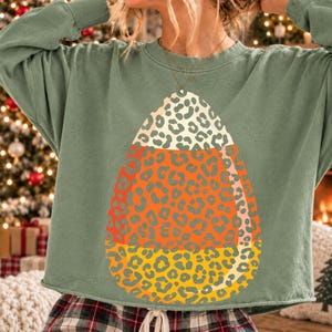 Könnte beinhalten: Ein langärmeliges Sweatshirt in Salbeigrün mit einer großen, zentralen Grafik in Form eines Zuckermais-Bonbons. Das Bonbon ist in drei horizontale Abschnitte unterteilt, die jeweils mit einem Leopardenmuster in Weiß, Orange und Gelb gefüllt sind.