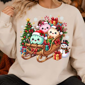 May include: A beige crewneck sweatshirt featuring a graphic print of three cartoon dumplings wearing Santa hats riding in a wooden sleigh filled with wrapped Christmas gifts, a decorated pine tree, candy canes, and a snowman standing nearby.