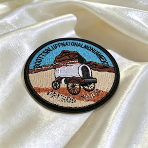 May include: Embroidered patch featuring a covered wagon and a mountain landscape. The patch is round with a black border and includes the text "SCOTTSBLUFF NATIONAL MONUMENT" and "OREGON TRAIL". The design incorporates shades of blue, brown, and tan.