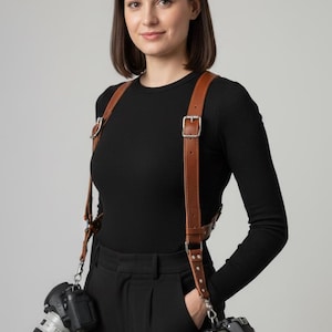 May include: A tan leather dual camera harness featuring adjustable straps with silver-toned metal buckles and hardware. The harness is designed to hold two cameras simultaneously, each attached to the lower ends of the leather straps via metal clips.
