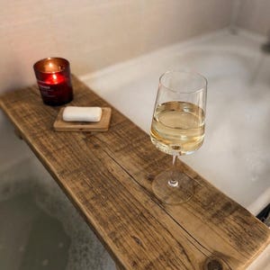 May include: A rustic wooden bathtub tray holds a lit red candle, a bar of soap on a wooden dish, and a glass of white wine. The tray spans the width of a white bathtub filled with bubbles.
