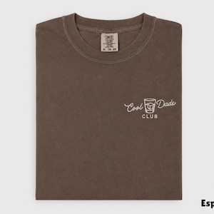 May include: Espresso-colored t-shirt with the words "Cool Dads Club" and a graphic of a cocktail glass. The shirt is folded and has a tag in the upper center. The shirt is a Comfort Colors brand.