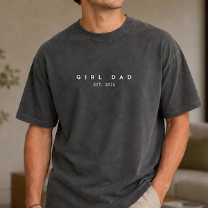 May include: A charcoal gray short-sleeve t-shirt with a crew neckline. The chest features white text that reads GIRL DAD and below it EST. 2026 in a smaller font. The fabric has a washed, vintage-style appearance with a relaxed fit.