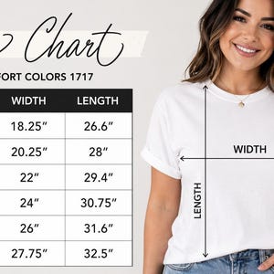 A woman is standing next to a size chart for a white t-shirt, smiling and posing for the camera.